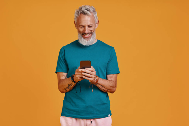 Happy senior man using smart phone and smiling while standing against orange background - Foto, imagen