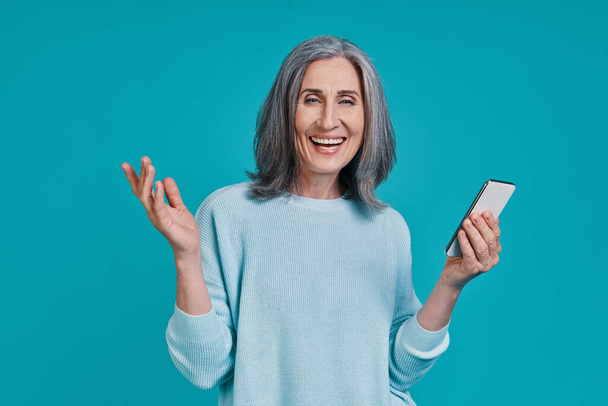 Mature beautiful woman using smart phone and smiling while standing against blue background - Fotó, kép