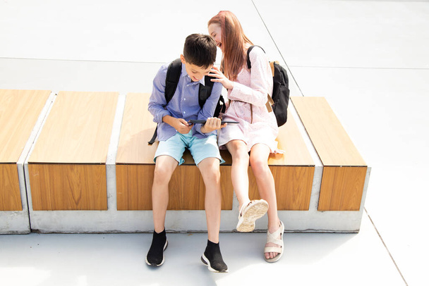 Schoolboy schoolgirl teenagers laugh have fun after lessons sitting on wooden bench in schoolyard, concrete background use tablet, concept of online education, modern technologies in kids life - Photo, image