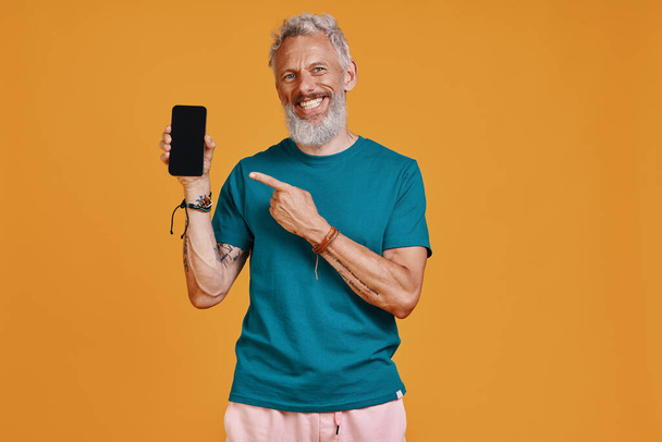 Happy senior man pointing smart phone and smiling while standing against orange background - Фото, зображення