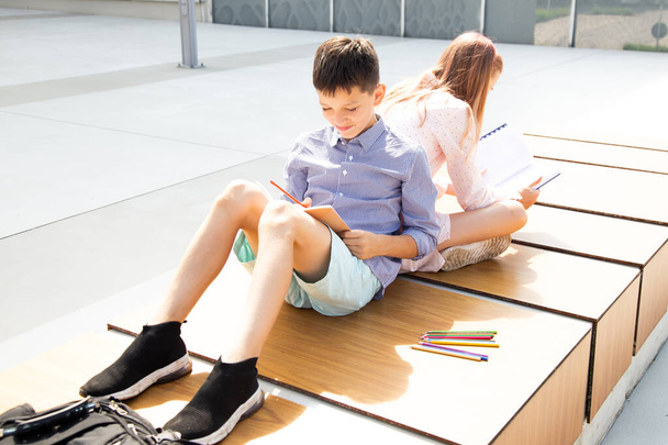 Öğrenciler okul bahçesinde sırt sırta otururken ödevlerini yaparlar. Gençlerin arkadaşlığı. - Fotoğraf, Görsel