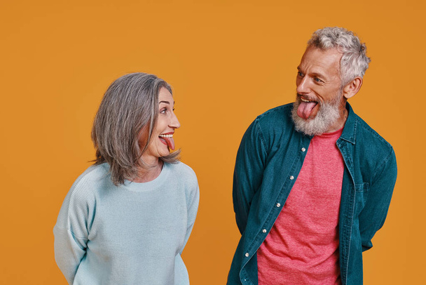Playful senior couple sticking out tongue while standing together against orange background - Foto, imagen
