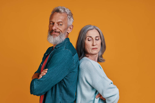 Displeased senior couple keeping arms crossed while standing together against orange background - Foto, Imagem