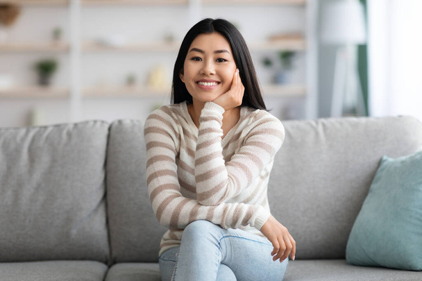 Портрет счастливой азиатки, сидящей на диване и улыбающейся перед камерой - Фото, изображение