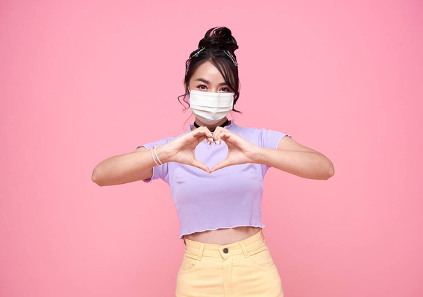 Щаслива жінка азіатського підлітка, яка показує міні-серце і носить маску обличчя, що захищає від Коровавіруса на рожевому тлі. охорона здоров "я. - Фото, зображення