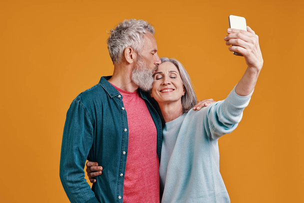 Beautiful senior couple using smart to take a selfie while standing together against orange background - 写真・画像