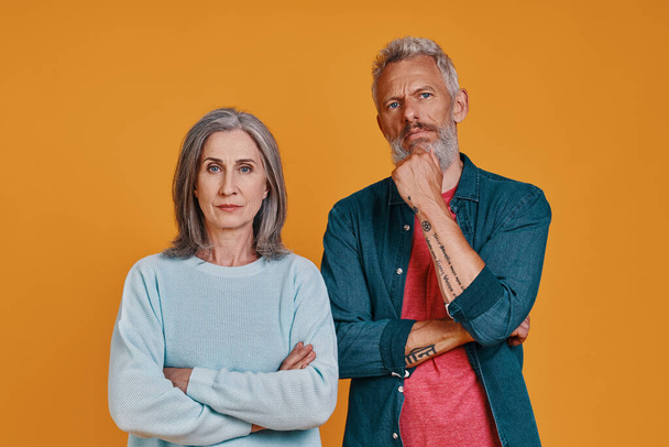 Thoughtful senior couple looking at camera while standing together against orange background - 写真・画像