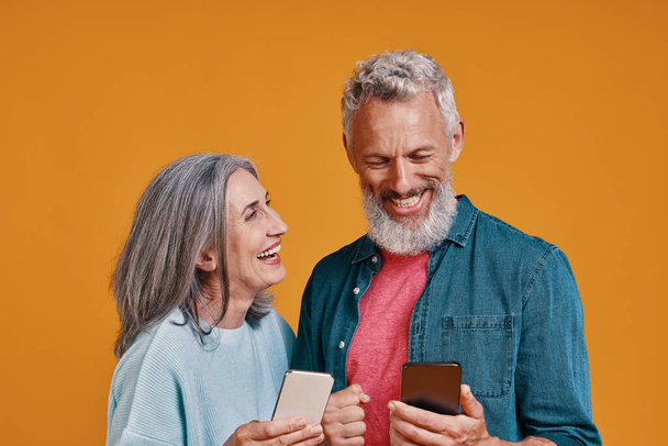 Happy senior couple smiling and using smart phones while standing together against orange background - Fotoğraf, Görsel