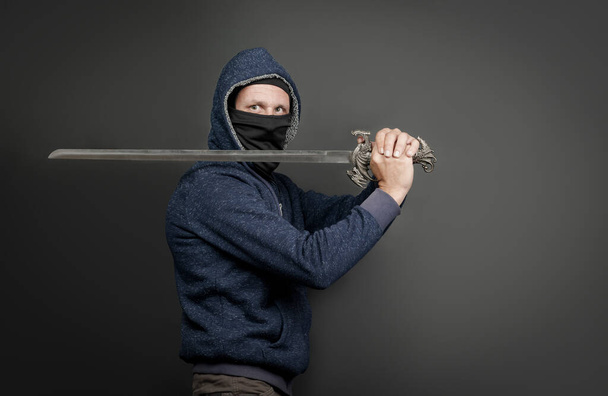 American Brutal man in a blue hoodie and black face mask with a Japanese sword. Portrait of a courageous warrior in jeans and a hood holding a big sword on the shoulder isolated on gray background - Fotoğraf, Görsel