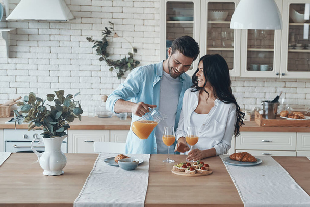 Handsome young man pouring orange juice while preparing breakfast together with his girlfriend - Foto, Imagem