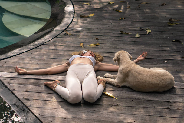 Young woman with curly long blonde hair and blue eyes in yoga asana and white dog next to blue swimming pool in Bali. - Photo, Image