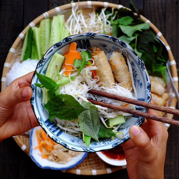 люди едят вьетнамскую вегетарианскую еду, рисовую лапшу, жареные весенние роллы с травами и соусом во время обеда, держат лапшу за руку на кулинарном фоне - Фото, изображение