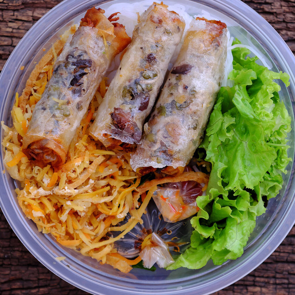 Widok z góry porcja z zamówienia online w plastikowej misce, wegańskie makaron ryżowy ze smażonymi sajgonkami i sałatką, sos, wegetariańskie jedzenie dla wietnamskiego wegetarianina na drewnianym tle - Zdjęcie, obraz