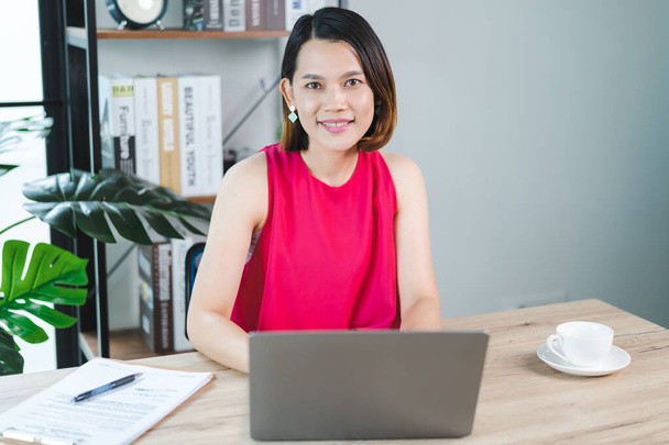 Wesoła, azjatycka bizneswoman w średnim wieku w luźnej, luźnej sukience pracująca w domu, sprawdzająca e-maile na laptopie, pisząca na papierze księgowym. Zdjęcie biznesowe - Zdjęcie, obraz