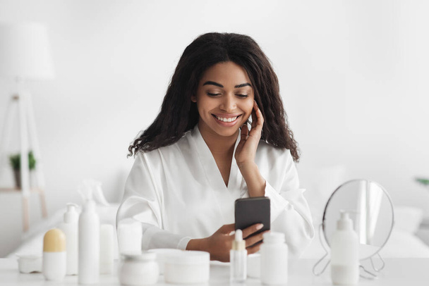 美容アプリケーションのコンセプト。アフリカ系アメリカ人女性の笑顔が寝室でスマホを使って白いバスローブを着て - 写真・画像