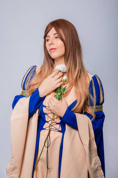 Portrait of an attractive woman with long hair in a medieval, fantasy, blue and beige dress with long, large sleeves, posing with white roses in her hands, isolated on a white background. - Foto, imagen