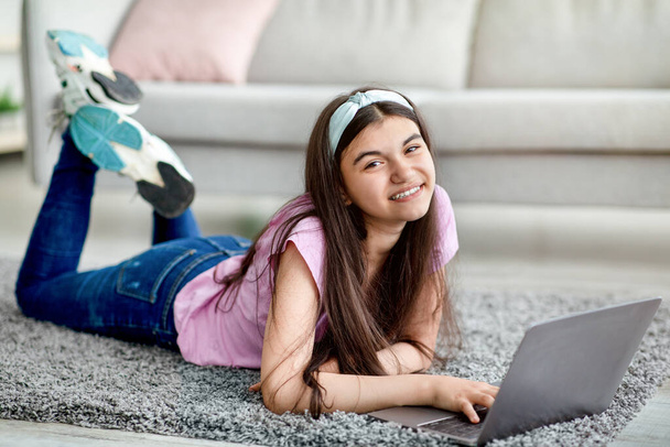 E-learning concept. Vrolijk adolescent meisje studeren online, liggend op de vloer met laptop thuis, volledige lengte - Foto, afbeelding