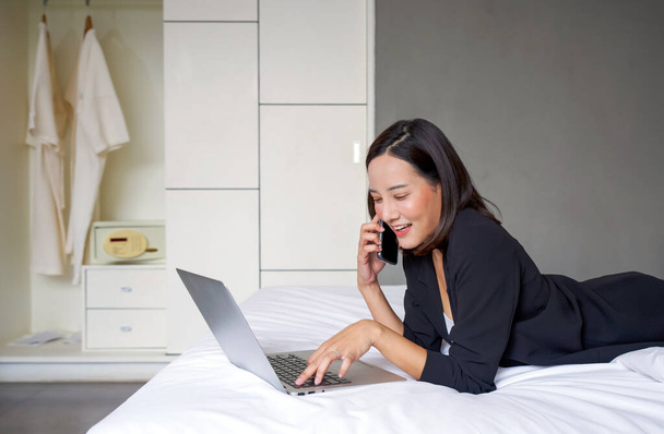 Una mujer asiática en traje negro usa la computadora portátil y el teléfono móvil para trabajar por la mañana en la cama. - Foto, imagen