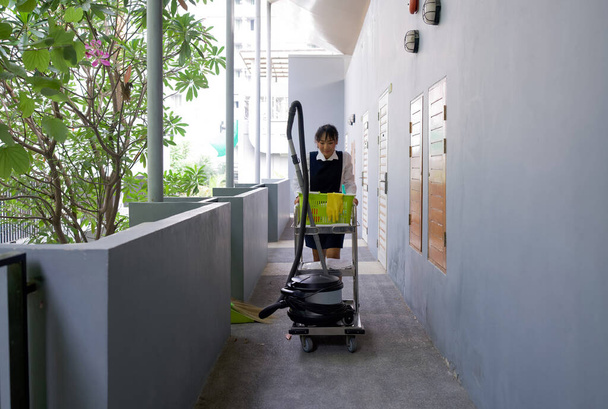 Ázsiai szobalány kék egyenruhában tolókocsi a folyosón a hotelszoba előtt. A szennyeskosár, az üvegtisztító, a gumikesztyű, a lábtörlő és a porszívó a kocsin van.. - Fotó, kép
