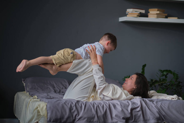 mom lying in bed have fun play with cute little son. Joyful young strong woman lifting on legs arms laughing little child , having fun together. - Photo, Image