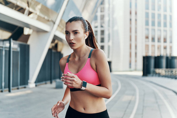 Beautiful young woman in sports clothing wearing headphones and smart watch while running outdoors - Фото, изображение