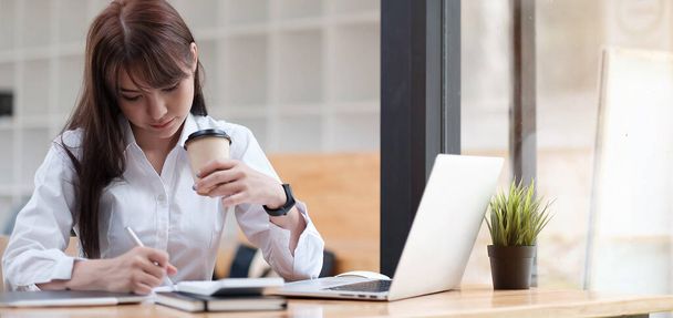 Bir kadın çay içiyor, ofiste yerde oturuyor, notlar yazıyor, önünde diz üstü bilgisayar açıyor. Merkez ofis kavramı. - Fotoğraf, Görsel