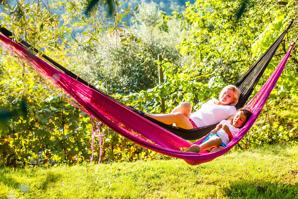 Close up portrait of a beautiful young mother and her five years old daughter laying down in a hammock playfully during a sunny summer day. Family lifestyle outdoors. - Photo, Image