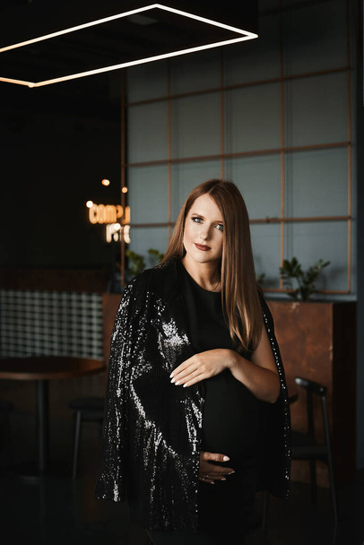 A young pregnant woman with bright makeup in a trendy black dress and fashionable shiny jacket in the dark interior - Zdjęcie, obraz