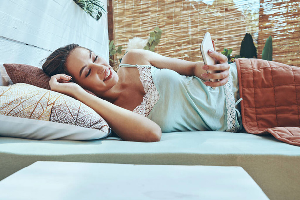Beautiful young smiling woman using mobile phone while lying down in domestic bed - Fotó, kép