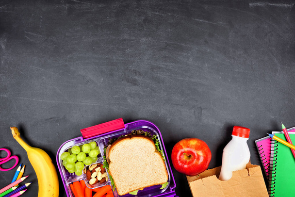 Healthy school lunch box and school supplies. Bottom border, top view on a chalkboard background. - Photo, Image
