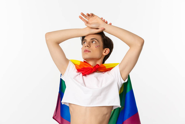 Schnappschuss eines sinnlichen androgynen Mannes mit bauchfreiem Oberteil und Regenbogenfahne, der mit erhobenen Händen in die linke obere Ecke blickt, weißer Hintergrund - Foto, Bild