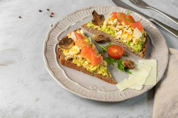 Healthy, nutritious sandwiches with a high protein content. Rye bread with scrambled eggs, avocado, salmon, baked mushrooms and parmesan. two toast with avocado, egg and salmon on a gray plate. Copy of the space - Photo, Image
