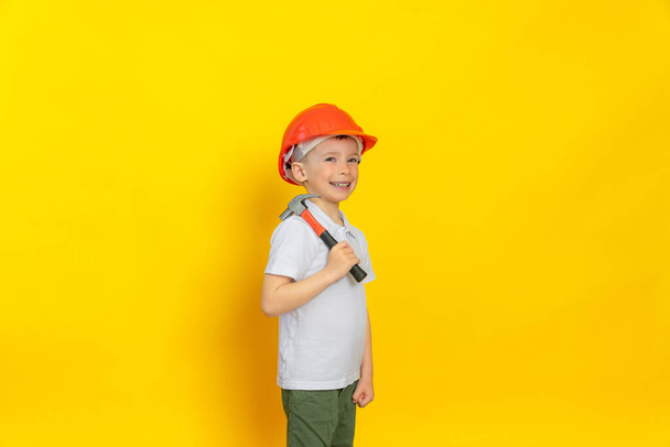 Cheerful and handsome child worker using a hammer construction tool. Dressed in a white T-shirt and a construction safety helmet. Yellow background. - Фото, изображение