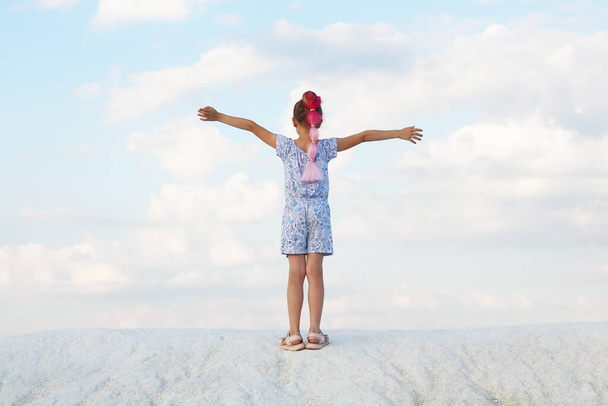 Nuori punatukkainen tyttö seisoo valkoisella vuorella sinistä taivasta vasten valkoisilla pilvillä kädet ylhäällä selkä valokuvaajalle - Valokuva, kuva
