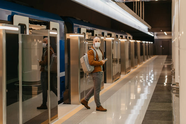 Egy férfi orvosi maszkban, hogy elkerülje a koronavírus terjedését, mobilt tart, miközben elhagyja a modern metrókocsit. Egy kopasz fickó sebészeti maszkban tartja a társadalmi távolságot.. - Fotó, kép
