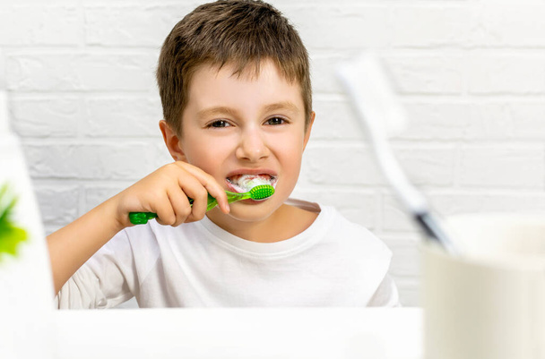 Portrait of happy cute kid boy in white pajamas brushes his teeth with a toothbrush and paste in the bathroom and looks at the camera. Dental health, daily hygiene and oral care for a child concept. - Fotó, kép