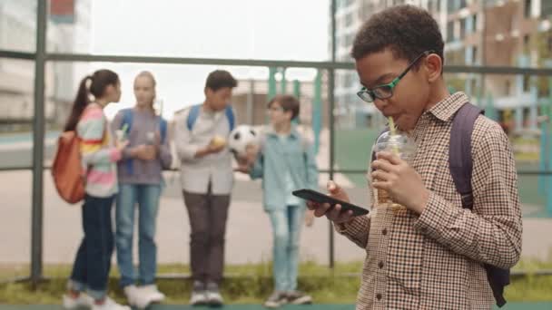 Medium slowmo πλάνο του μικτού σχολείου αγόρι με φρέσκια λεμονάδα και smartphone στέκεται στην παιδική χαρά ξοδεύοντας χρόνο με τους συμμαθητές μετά το σχολείο - Πλάνα, βίντεο