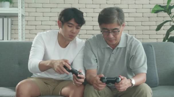 Asian family with son teaching father play video game on sofa at home, elderly and young man two generations competition enjoying joystick with entertainment and learning, lifestyles concept. - Filmagem, Vídeo