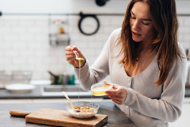 Young woman in the kitchen adds honey to granola - Photo, Image
