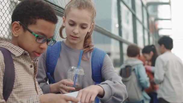 Közepes lassítású felvétel többnemzetiségű fiatalkorú iskolás fiúról és lányról, akik otthoni feladatokról beszélgetnek, miközben a szabadtéri sportpályán állnak. - Felvétel, videó