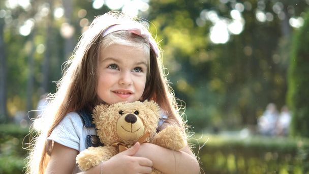 Happy little preeschool girl playing with her favorite teddy bear toy outdoors in summer park. - 写真・画像
