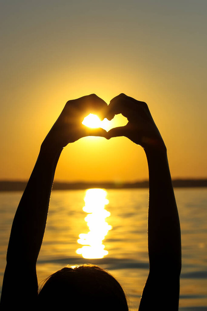 Silhouette des mains de la femme en forme de cœur au coucher du soleil contre un ciel jaune-orange, des rayons de soleil et un soleil affiché à l'intérieur. Symbole d'amour. Vacances en bord de mer, océan, lac, rivière en été. Bord de mer, côte. - Photo, image
