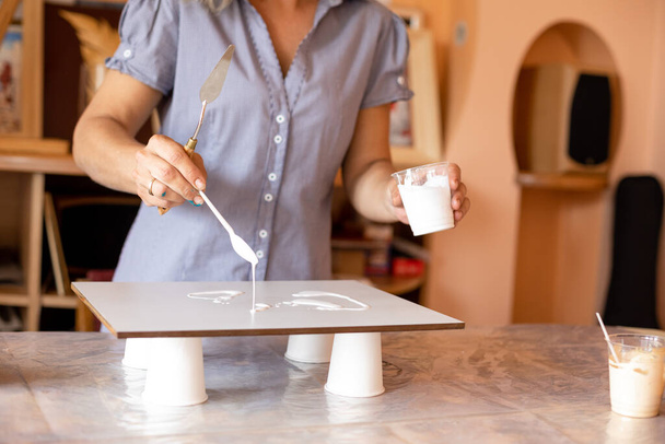 Artist holds cup of white paint in her left , and with her right hand she applies paint to surface to create white background. Creativity and design. Freedom and inspiration. Hobbies and crafts. - Foto, afbeelding