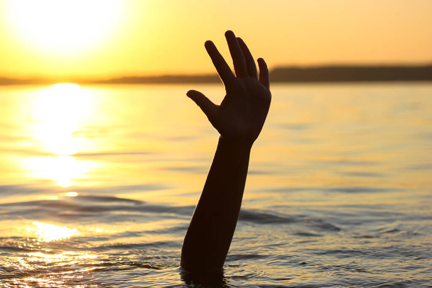 Mies hukkuu veteen auringonlaskun aikaan. Käsi kurkistaa veden alta. Hukkuva ihminen tarvitsee apua, pelastusta. Riski, hengenvaara meressä, joessa, meressä. Kuolema, SOS-käsite. Hukkuva uhri. - Valokuva, kuva