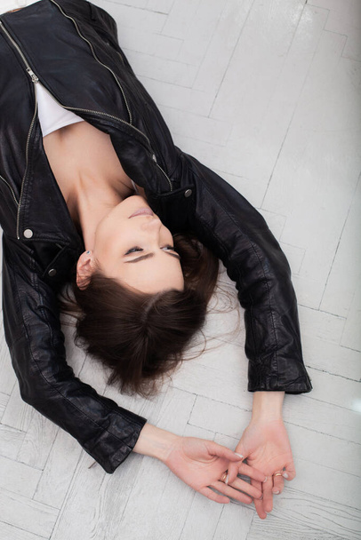 a brunette girl in black leather jacket lying on a white parquet floor background with her hands on the floor over head - Zdjęcie, obraz