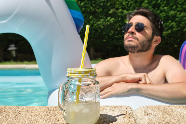 水泳プールのユニコーンインフレータブルリングにサングラスをかけたレモネードドリンクと若い男。夏のコンセプト. - 写真・画像