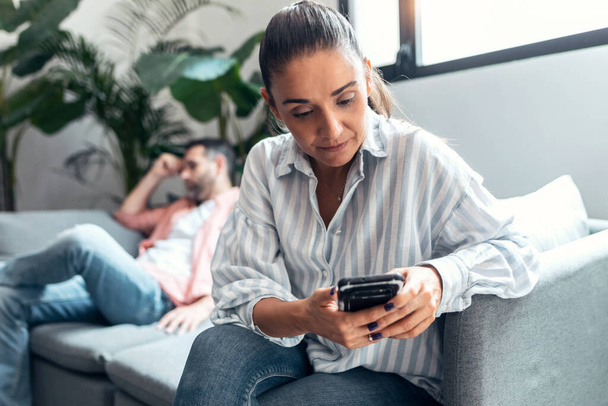 Shot of angry young couple sitting on couch together and looking to opposite sides while she using mobile phone at home. - Foto, imagen