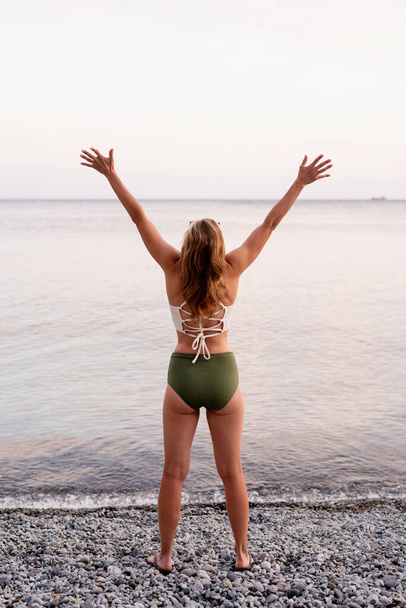 Wakacje. Widok z tyłu młodej kobiety stojącej na kamienistej plaży z wyciągniętymi ramionami - Zdjęcie, obraz