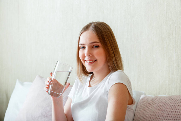 Smile young woman drinks glass of pure water in morning after waking up in her bed room. Happy teen girl maintains water balance for body health by drinking clean water. - 写真・画像