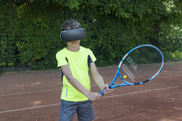 Хлопчик в окулярах віртуальної реальності тримає ракетку і грає в теніс з віртуальним тренером на наземному корті. Навчальний процес у новій реальності
. - Фото, зображення